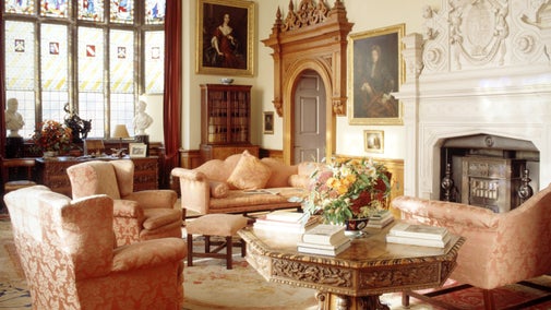 Historic sitting room with pink upholstered furniture, an ornate fireplace, stained-glass windows, and a carved wooden table topped with books and flowers.